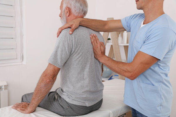 Un homme âgé assis sur une table d'examen reçoit une séance d'ostéopathie de la part d'un professionnel de santé en tenue médicale bleue, qui pose ses mains sur le dos et l'épaule du patient dans un cabinet lumineux.