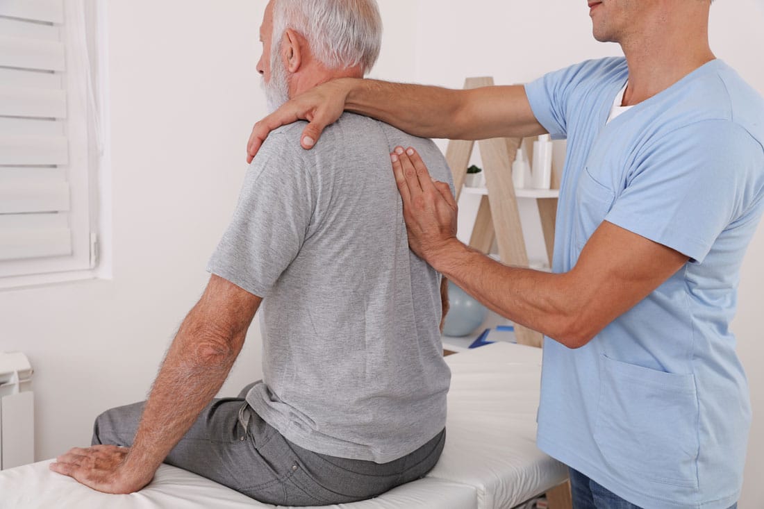 Un homme âgé assis sur une table d'examen reçoit une séance d'ostéopathie de la part d'un professionnel de santé en tenue médicale bleue, qui pose ses mains sur le dos et l'épaule du patient dans un cabinet lumineux.