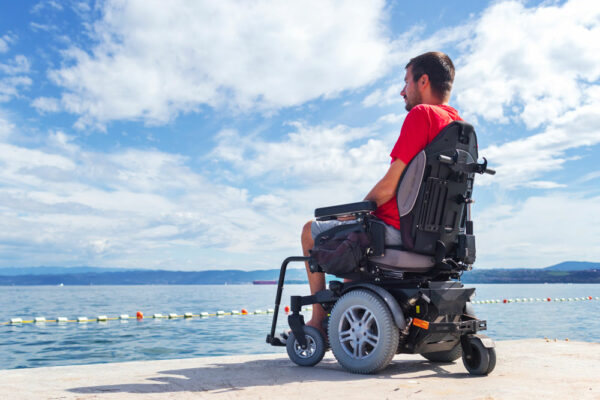 fauteuil-electrique-pris-en-charge Un homme assis dans un fauteuil roulant électrique regarde l'eau tout en étant assis sur une jetée ou un quai. Il porte un t-shirt rouge et un short gris, sous un ciel bleu partiellement nuageux. Le paysage en arrière-plan montre un plan d'eau et des montagnes ou collines au loin.