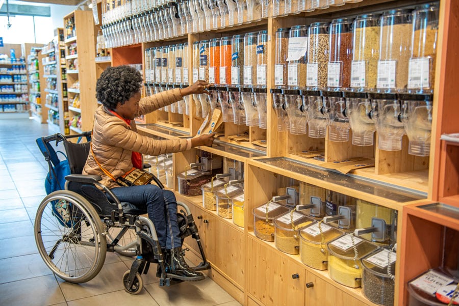 Une femme en fauteuil roulant se sert en vrac dans un magasin bio. Elle remplit un sac en papier en utilisant un distributeur transparent situé sur une étagère en bois garnie de divers produits alimentaires en vrac comme des céréales, des graines et des légumineuses, illustrant l'accessibilité et la consommation responsable.