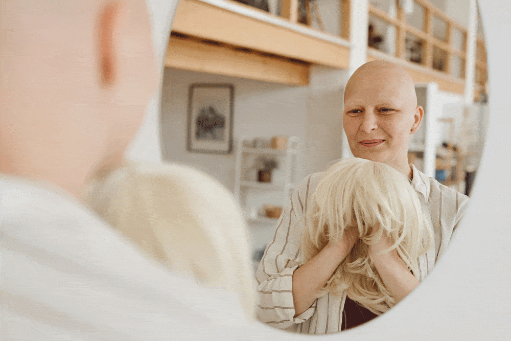 Une femme chauve sourit en se regardant dans un miroir tout en tenant une perruque blonde devant elle. La scène se déroule dans un intérieur lumineux et moderne, et l'expression de la femme semble confiante et sereine.